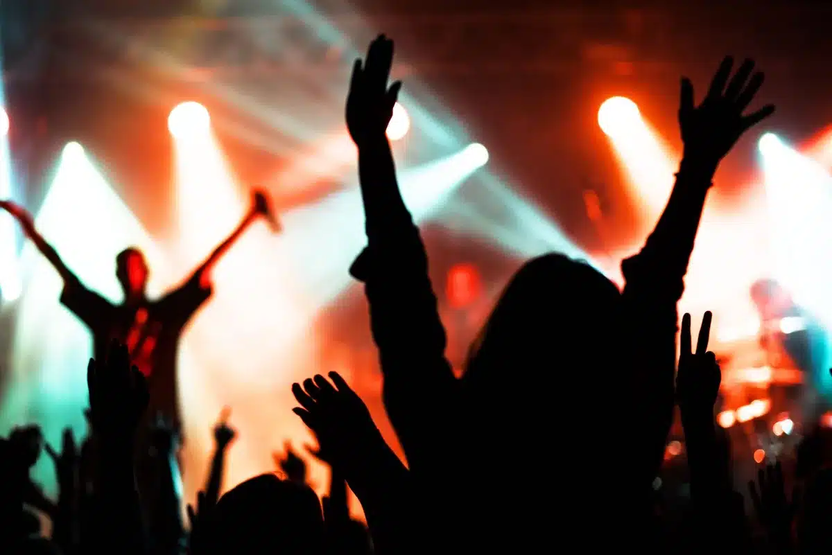 Crowd of music fans with raised hands at an outdoor concert stage with colorful lights
